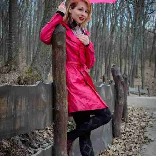 Young stylish female wearing trendy pink raincoat leaning on wooden fence while holding umbrella overhead while standing on walkway in autumn park
