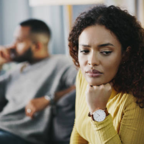 Black woman in deep thought contemplating relationship decisions while partner sits distant on phone in background